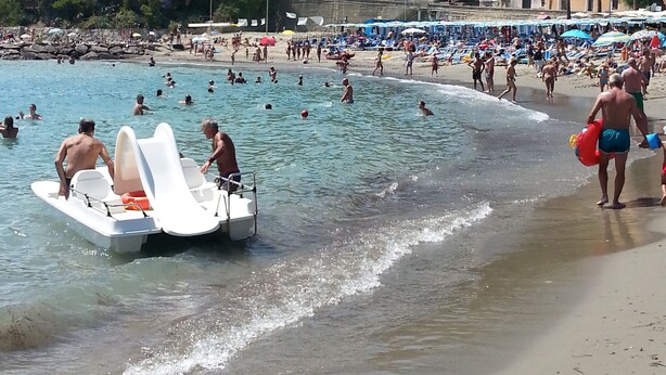 A beach with people swimming and sunbathing, a boat on the shore, and buildings in the background.