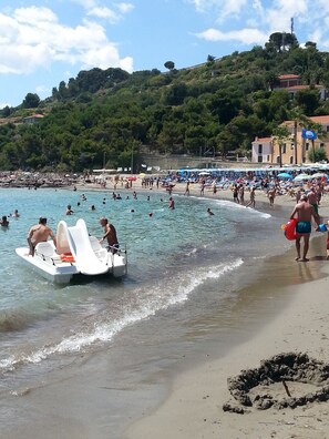 Una playa con gente nadando y tomando el sol, un bote en la orilla y edificios al fondo.
