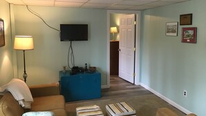 A living room with a flat-screen TV mounted on the wall, a blue ottoman, a beige sofa, and a lamp. - Room in Penn State area (State College)
