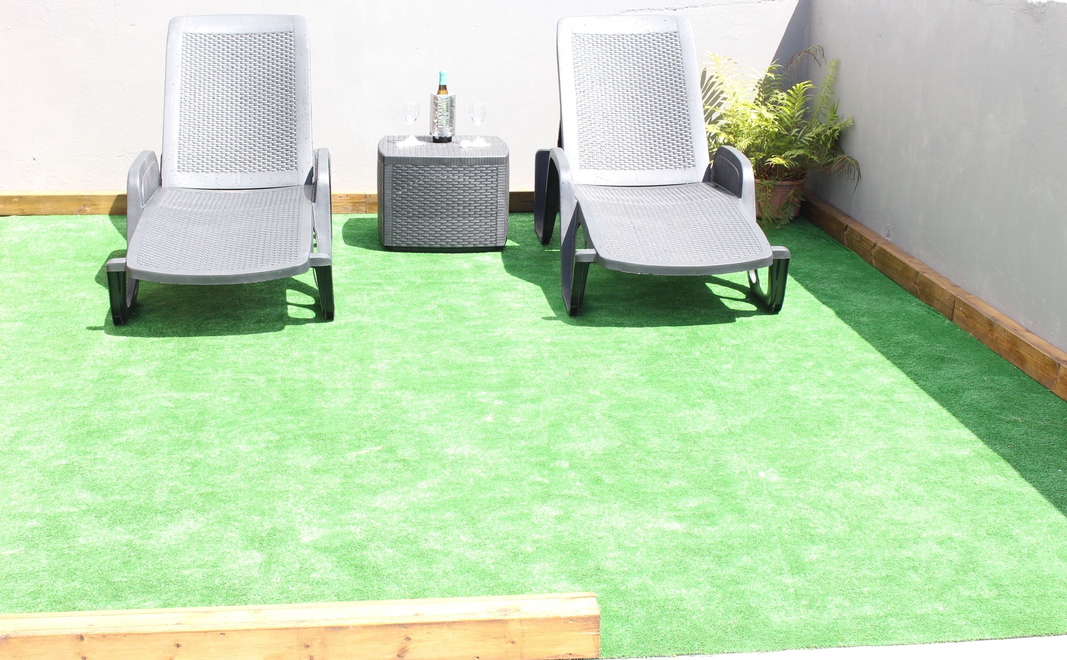 Two grey wicker chairs with metal legs on a green artificial turf, a small round table with a bottle and glass, and a potted plant against a wall.