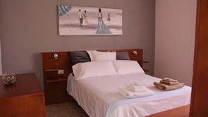 Ein ordentlich bezogenes Bett mit weißen Leinen und einer Streifen-Decke, ein Nachttisch aus Holz mit einer Schale Nüsse aus Glas und ein Bild von drei Personen am Strand, das über dem Bett hängt.