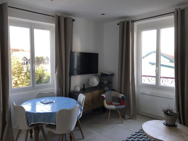 Une pièce comprenant une table à manger, des chaises, un téléviseur fixé au mur et une fenêtre avec des rideaux.