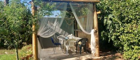 A covered outdoor seating area with a table and chairs, surrounded by lush greenery.