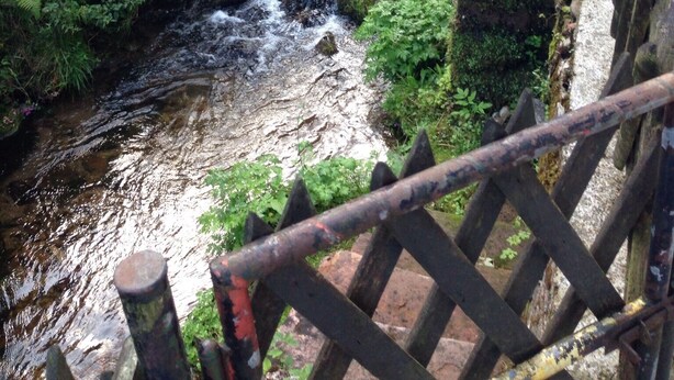 Un ponte di pietra con ringhiera in metallo che sovrasta un ruscello in una zona verde e rigogliosa.