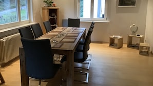 Una sala da pranzo con un lungo tavolo di legno apparecchiato per sei persone, circondato da sedie. Due finestre lasciano entrare luce naturale, un termosifone a sinistra e una piccola mensola con oggetti decorativi.