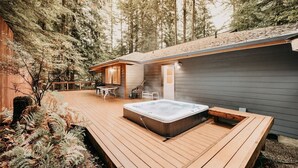 A wooden deck with a hot tub, a small wooden structure with a table and chairs, and a forested backdrop. - 29sl - Hot Tub - Sauna - Bbq - Shuffle Board - Sleeps 11 3 Bedroom Home by RedAwning (Deming)