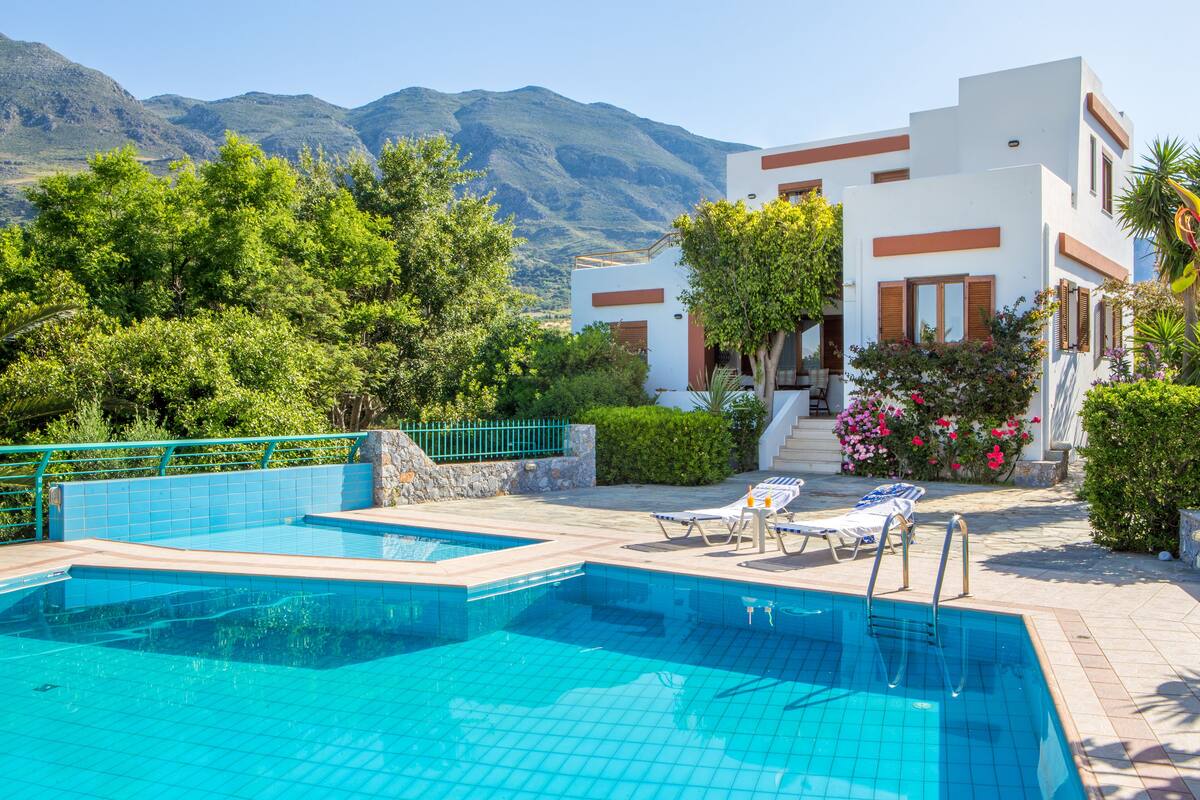 Une piscine divisée en deux zones, entourée d’un jardin aménagé et bordée d’un bâtiment blanc agrémenté de touches de couleur marron.