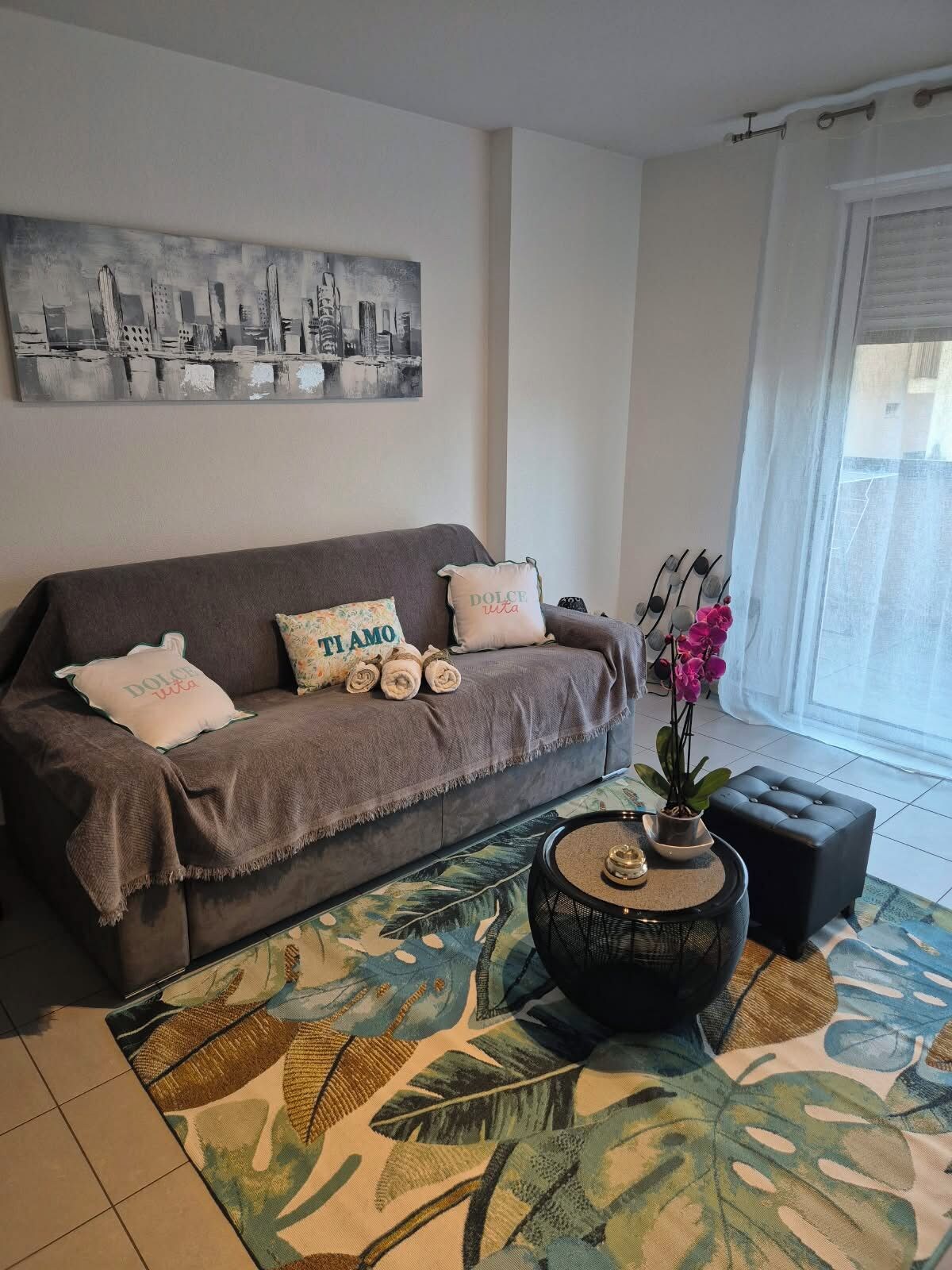 A living room with a sofa, decorative pillows, a cityscape painting, a potted plant, and a patterned rug.