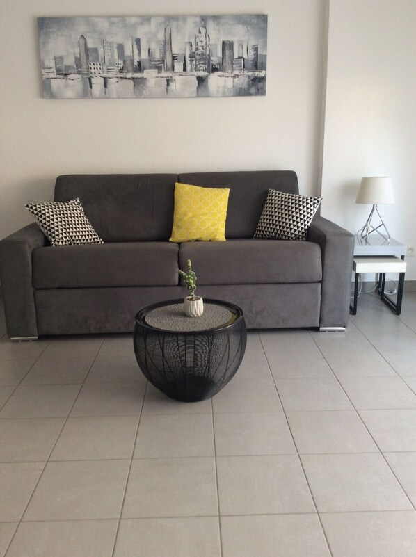 Una sala de estar moderna con un sofá gris, cojines estampados, una mesa redonda de centro con una planta y un cuadro de un paisaje urbano en la pared.