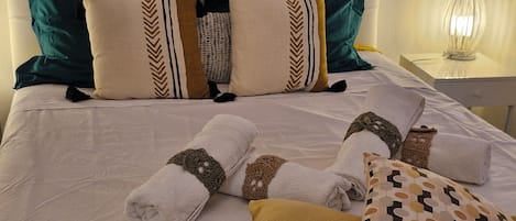 A neatly made bed with rolled towels, pillows with geometric patterns, and a bedside lamp.