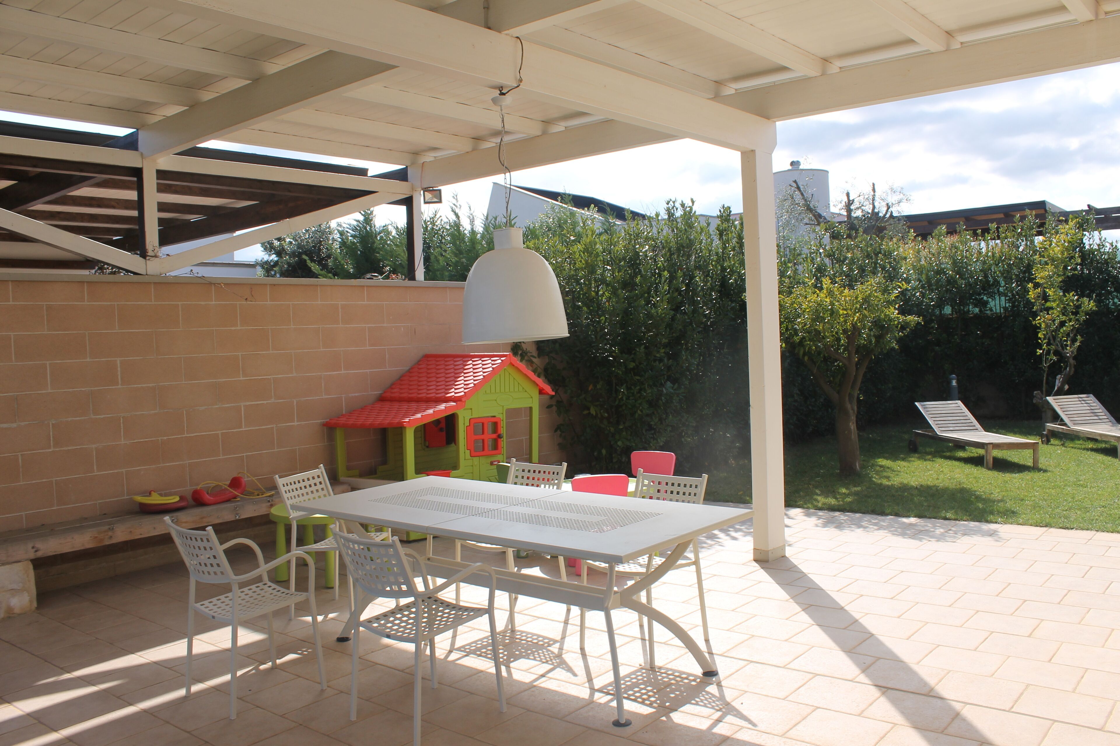 An outdoor patio with a white table and chairs, a hanging light fixture, and a small playhouse in the background.