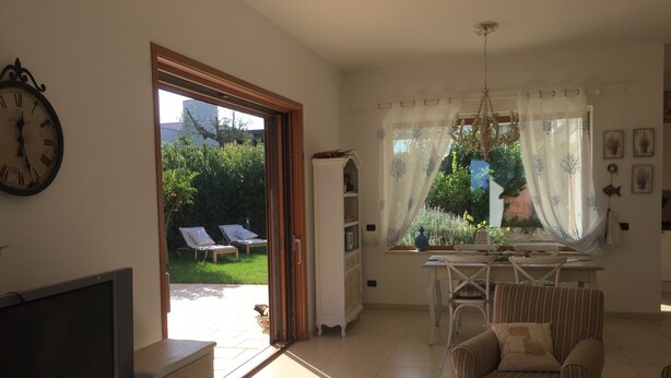 Una sala de estar con televisor, sofá, zona de comedor con mesa y sillas, y vista al jardín a través de un ventanal grande.