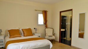 Una camera da letto con un letto, comodini, una sedia, una finestra con tende, una porta che conduce al bagno e uno specchio appeso al muro.