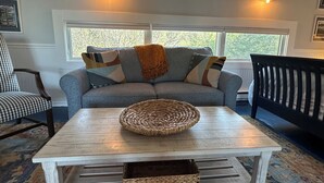 Ein gemütliches Wohnzimmer mit einem grauen Sofa, einem weißen Couchtisch und einer schwarzen Bank. Zwei Wandleuchten und ein Fenster mit Blick auf Bäume.