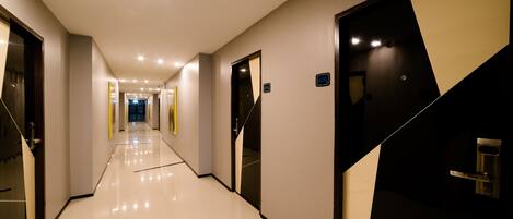 A hotel corridor with a modern design, featuring a reflective floor, geometric wall panels, and a series of doors leading to rooms.