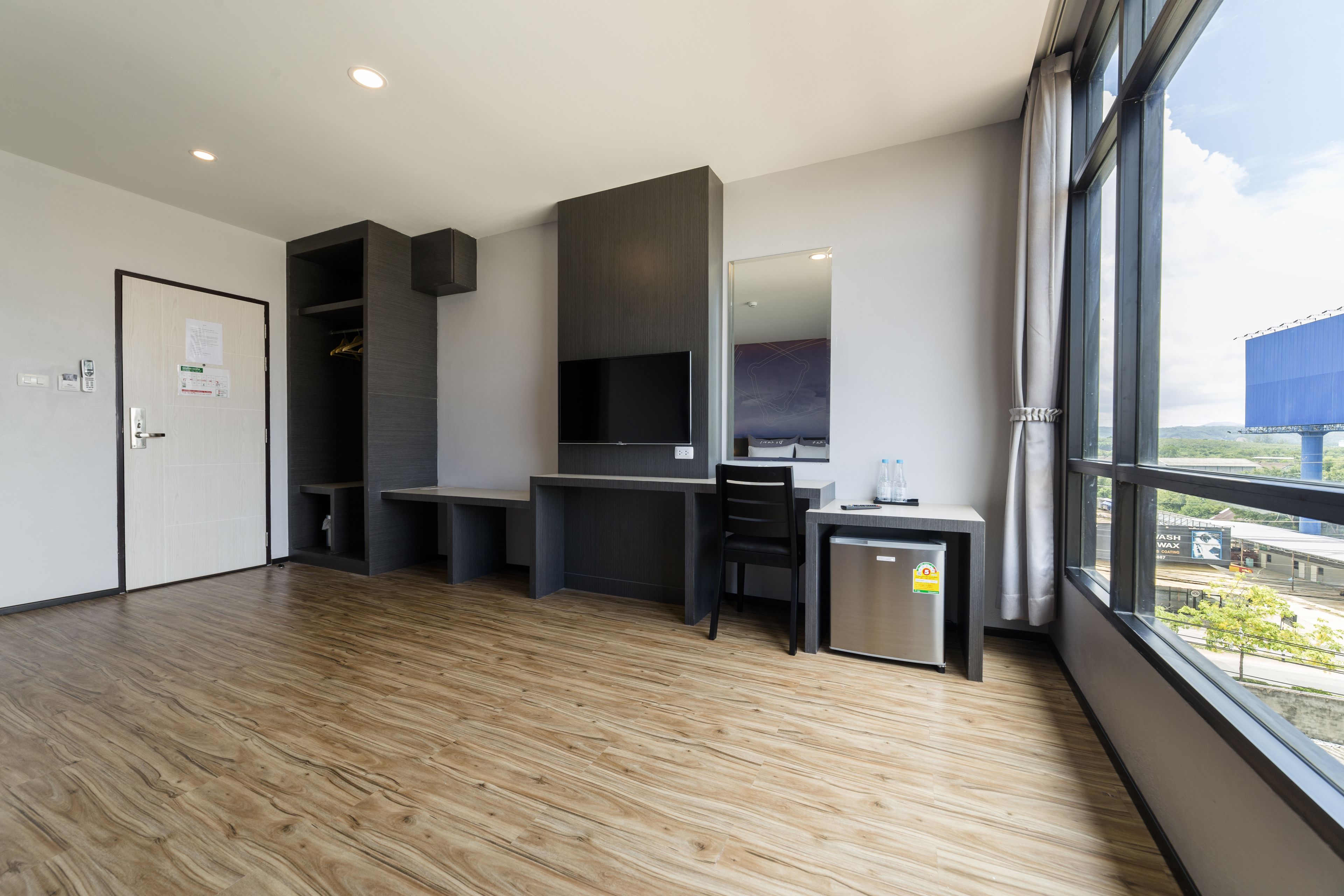 Una habitación de hotel moderna con piso de madera, un escritorio con computadora, un televisor y un refrigerador pequeño.
