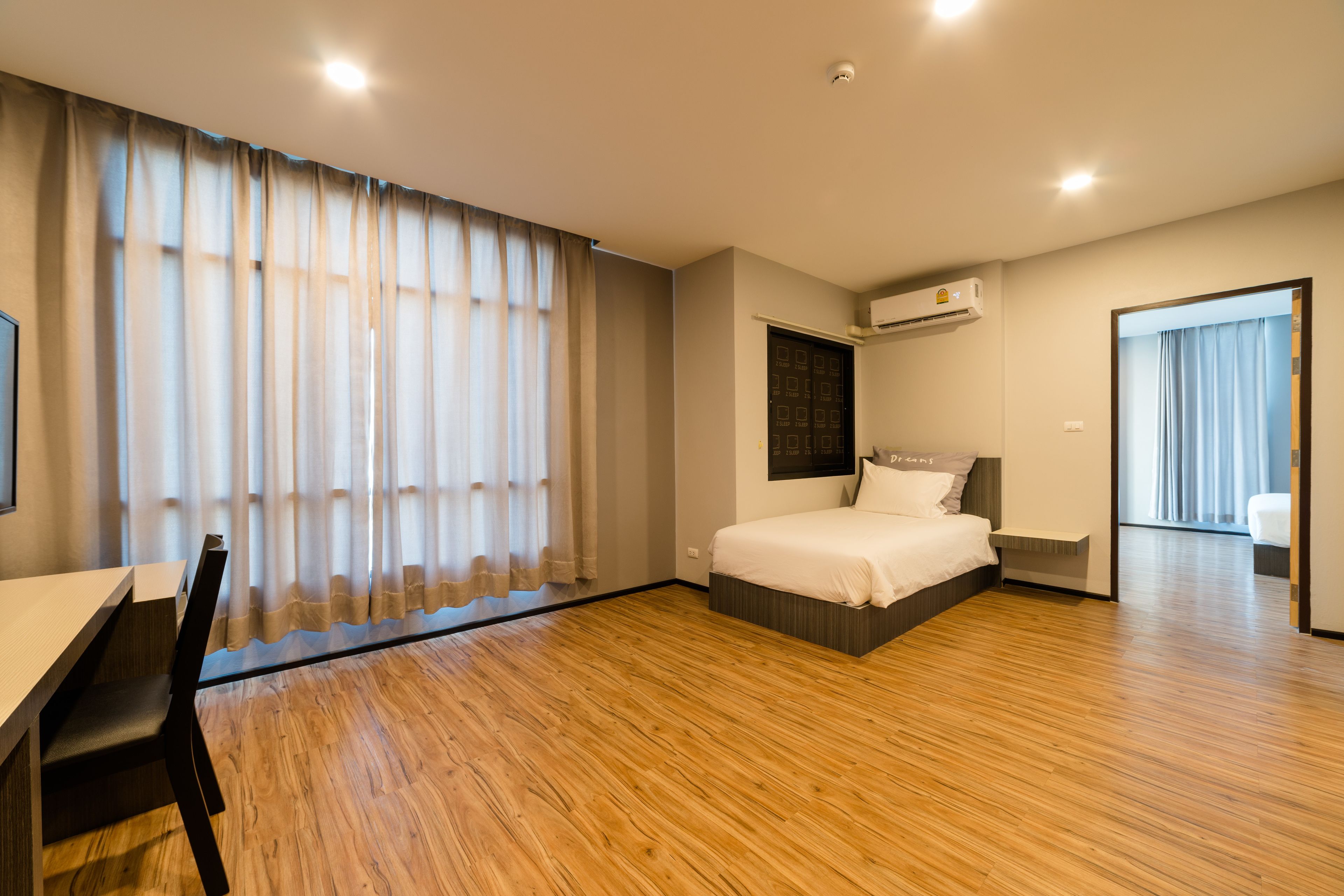 Una habitación de hotel moderna con piso de madera, una cama con cabecera, un escritorio con silla y un ventanal con cortinas.