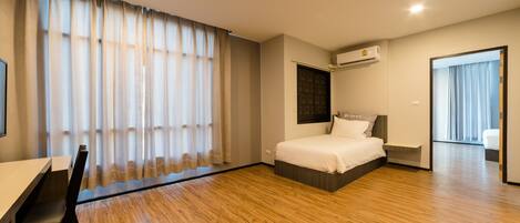 Una habitación de hotel moderna con piso de madera, una cama con cabecera, un escritorio con silla y un ventanal con cortinas.