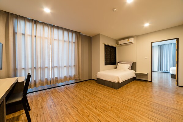 Una habitación de hotel moderna con piso de madera, una cama con cabecera, un escritorio con silla y un ventanal con cortinas.