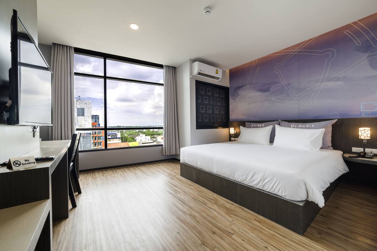 A modern hotel room with a large bed, a desk, and a view of the city through large windows.