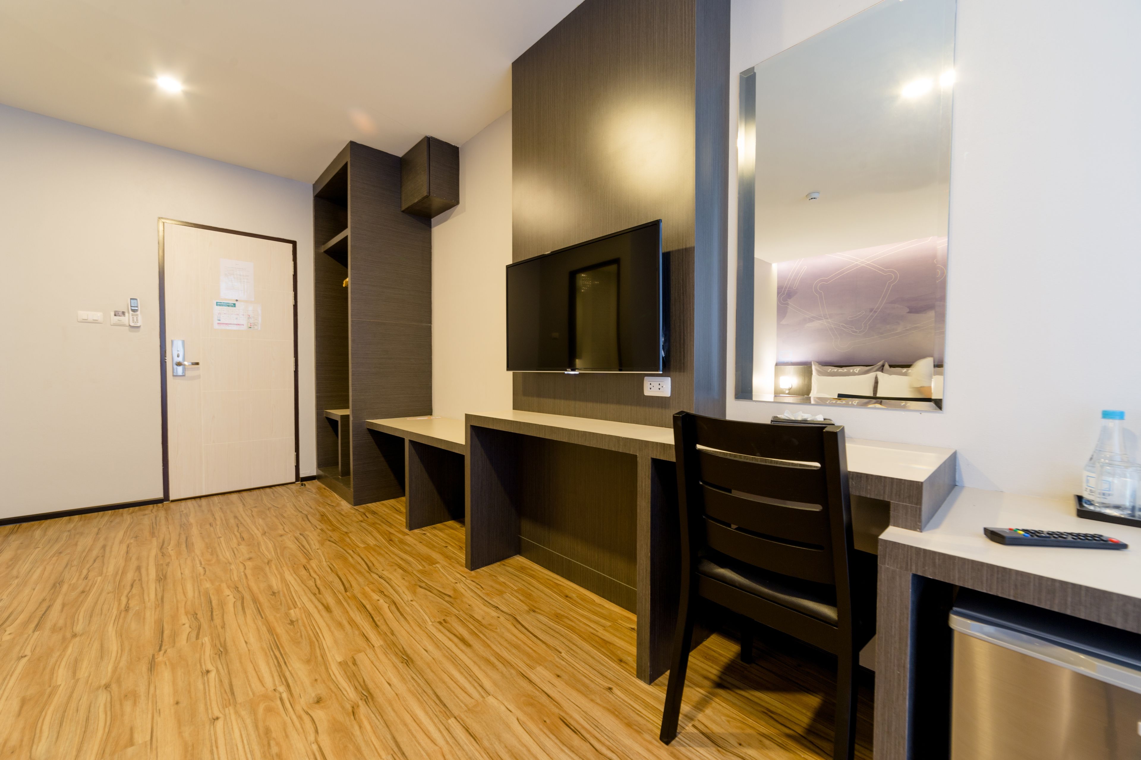 A modern hotel room with a wooden floor, a desk with a chair, a flat-screen TV mounted on the wall, a mini-fridge, and a water bottle on the counter.