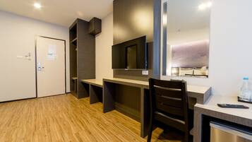 A modern hotel room with a wooden floor, a desk with a chair, a flat-screen TV mounted on the wall, a mini-fridge, and a water bottle on the counter.