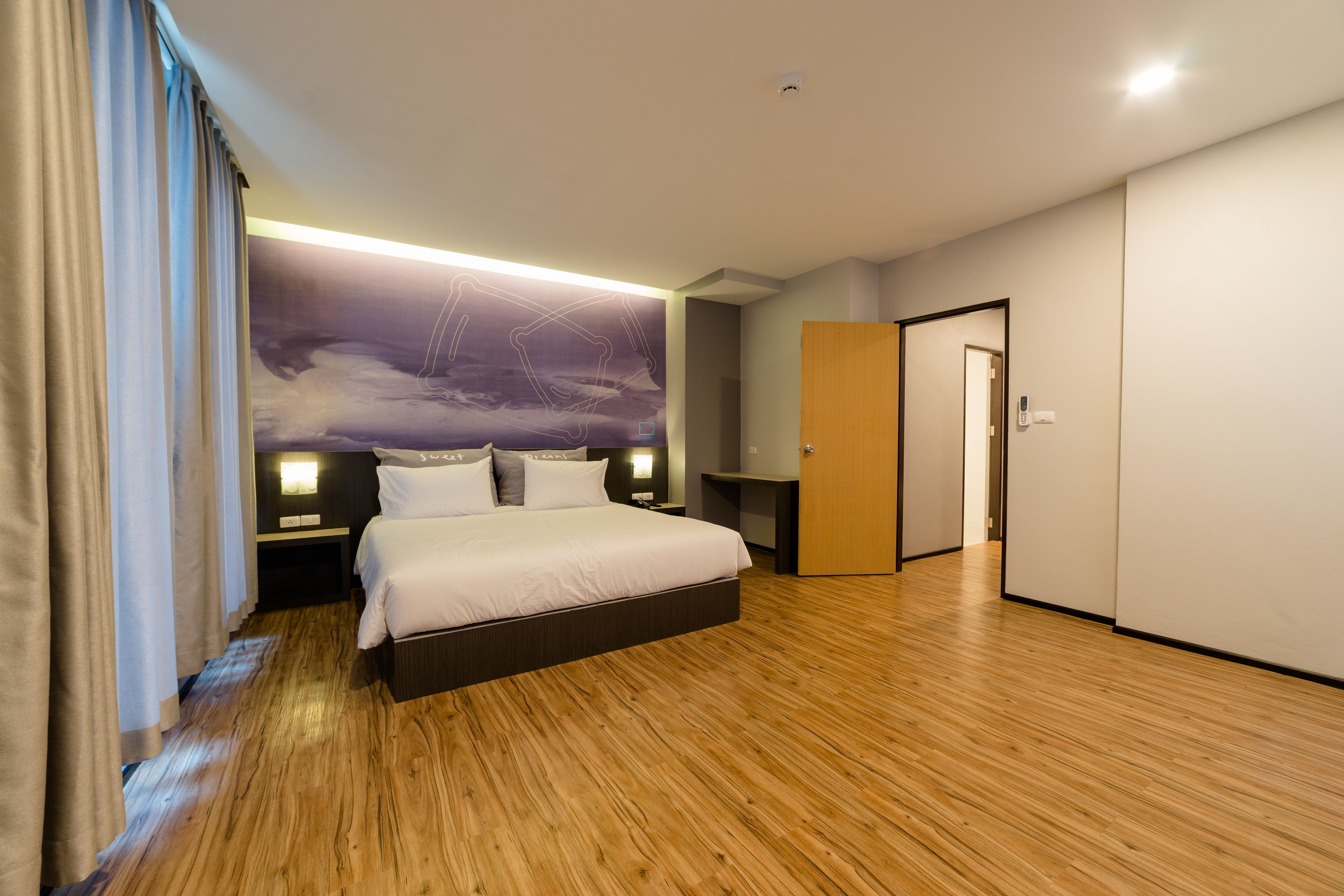 A modern hotel room with a large bed, wooden flooring, a nightstand, a wall-mounted lamp, and a door leading to another room.