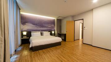 A modern hotel room with a large bed, wooden flooring, a nightstand, a wall-mounted lamp, and a door leading to another room.