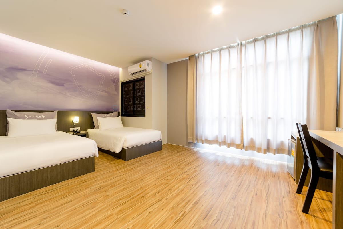 A hotel room with two beds, a desk, a chair, a wall-mounted air conditioner, and a large window with curtains.