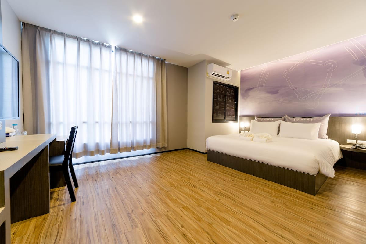 A hotel room with a wooden floor, a bed with white bedding, a desk with a chair, and a large window with curtains.