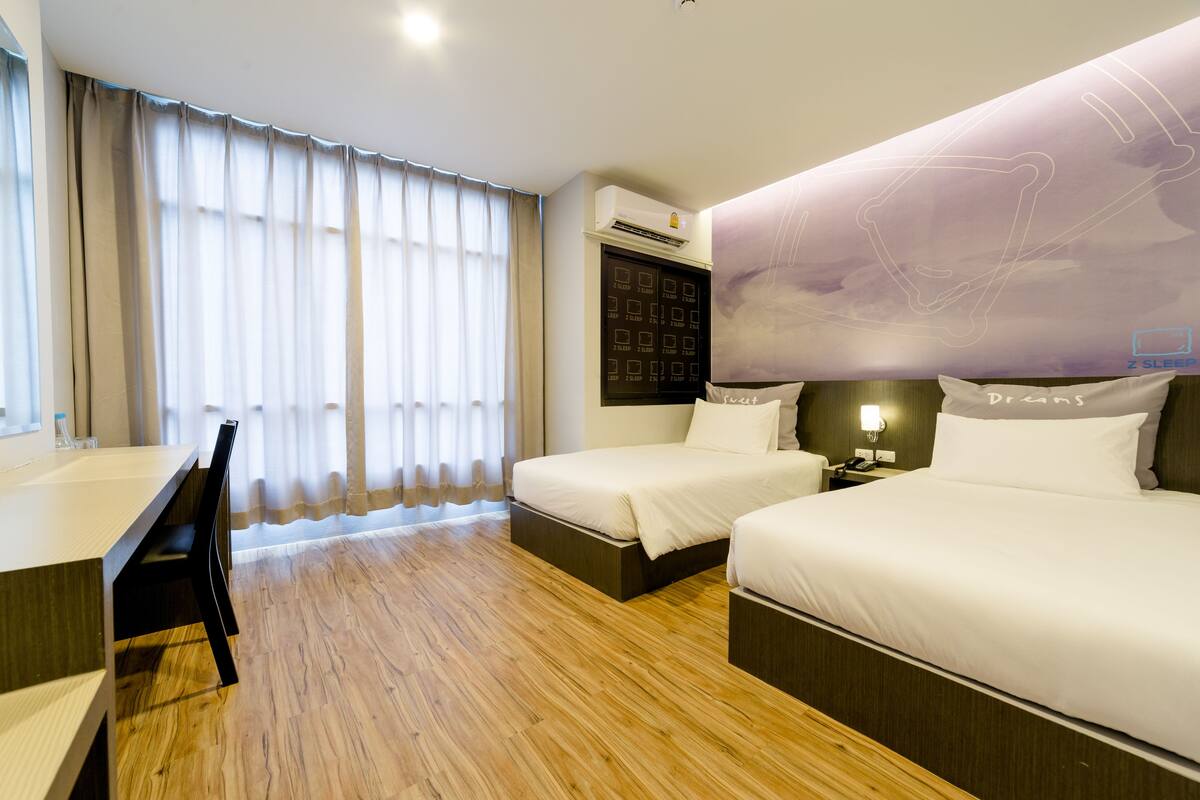 A hotel room with two beds, a desk, and a large window with curtains.