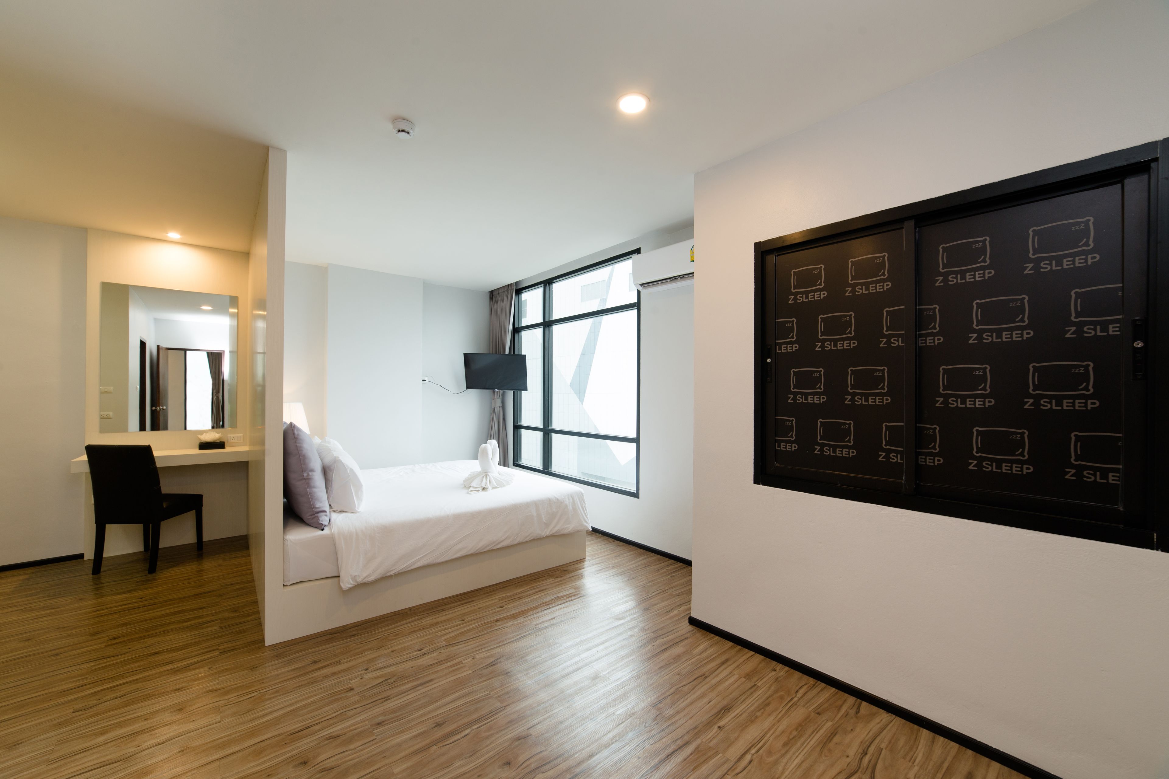 A modern hotel room with a bed, a desk, a chair, a flat-screen TV, and a wall with multiple beds labeled 'Z SLEEP'.