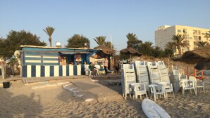 Un café frente a la playa con terraza, una tabla de surf y sillas apiladas.