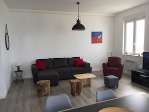 A modern living room with a dark grey sofa, red cushions, a wooden coffee table, a red armchair, and a small blue ottoman. There is a painting on the wall and a black pendant light above the sofa.