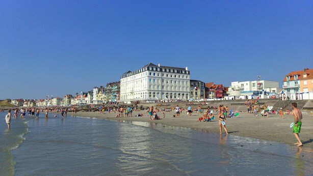 Een strand met mensen, een groot wit gebouw en kleurrijke huizen op de achtergrond.