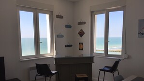 Een kamer met twee ramen met uitzicht op de zee, een kleine bar en een eettafel met stoelen.