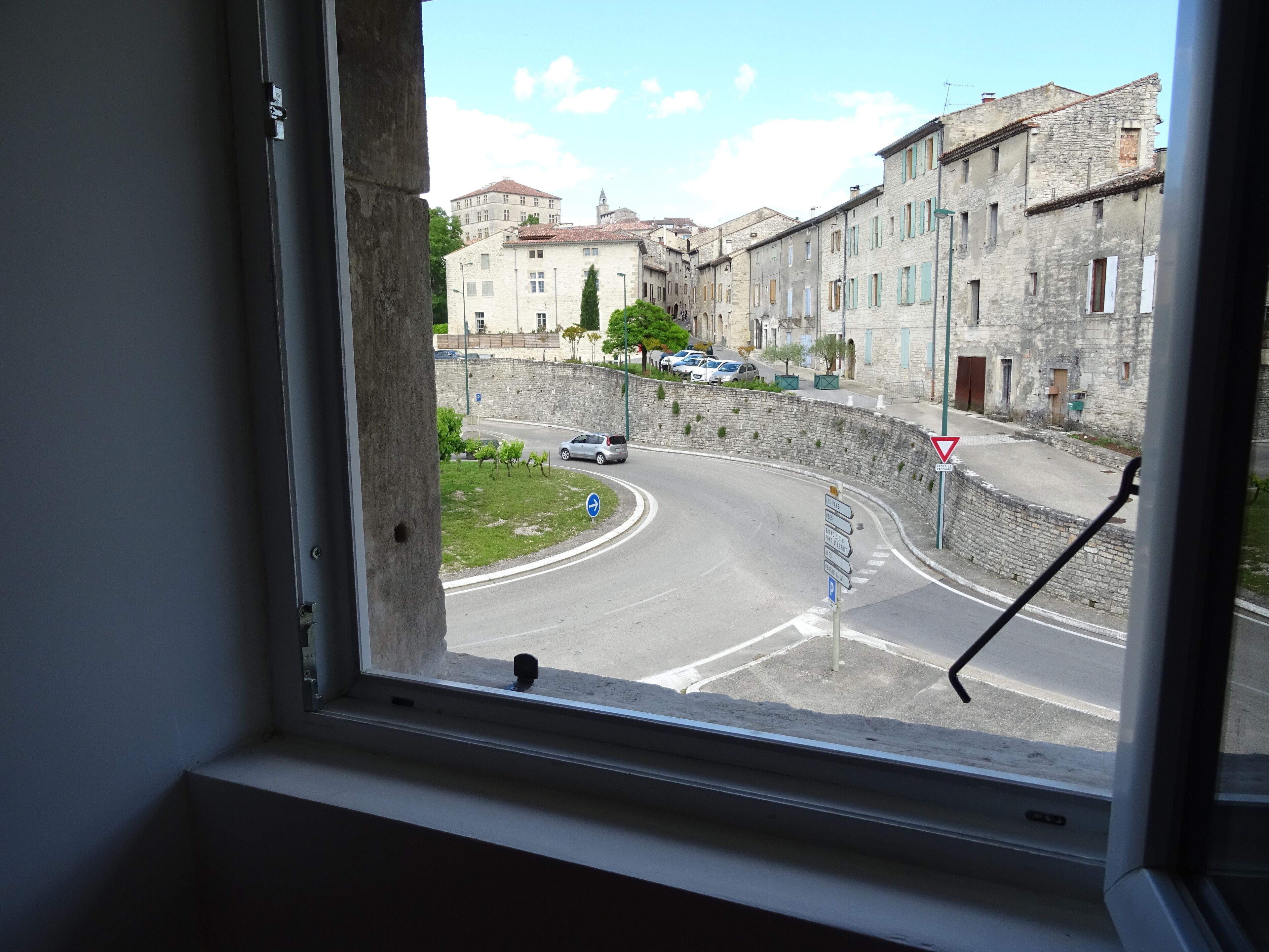 Vista dalla finestra di un edificio con facciate in pietra, una rotonda e auto parcheggiate.