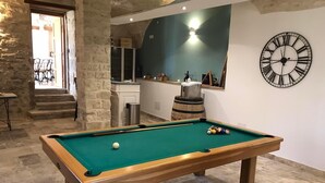 Una mesa de billar con bolas, un reloj y una zona de bar en una habitación con paredes de piedra y una pared azul de acento.