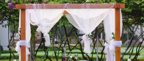 Un pabellón con cortinas blancas y decoración floral, rodeado por un jardín con una mesa dispuesta para un evento.
