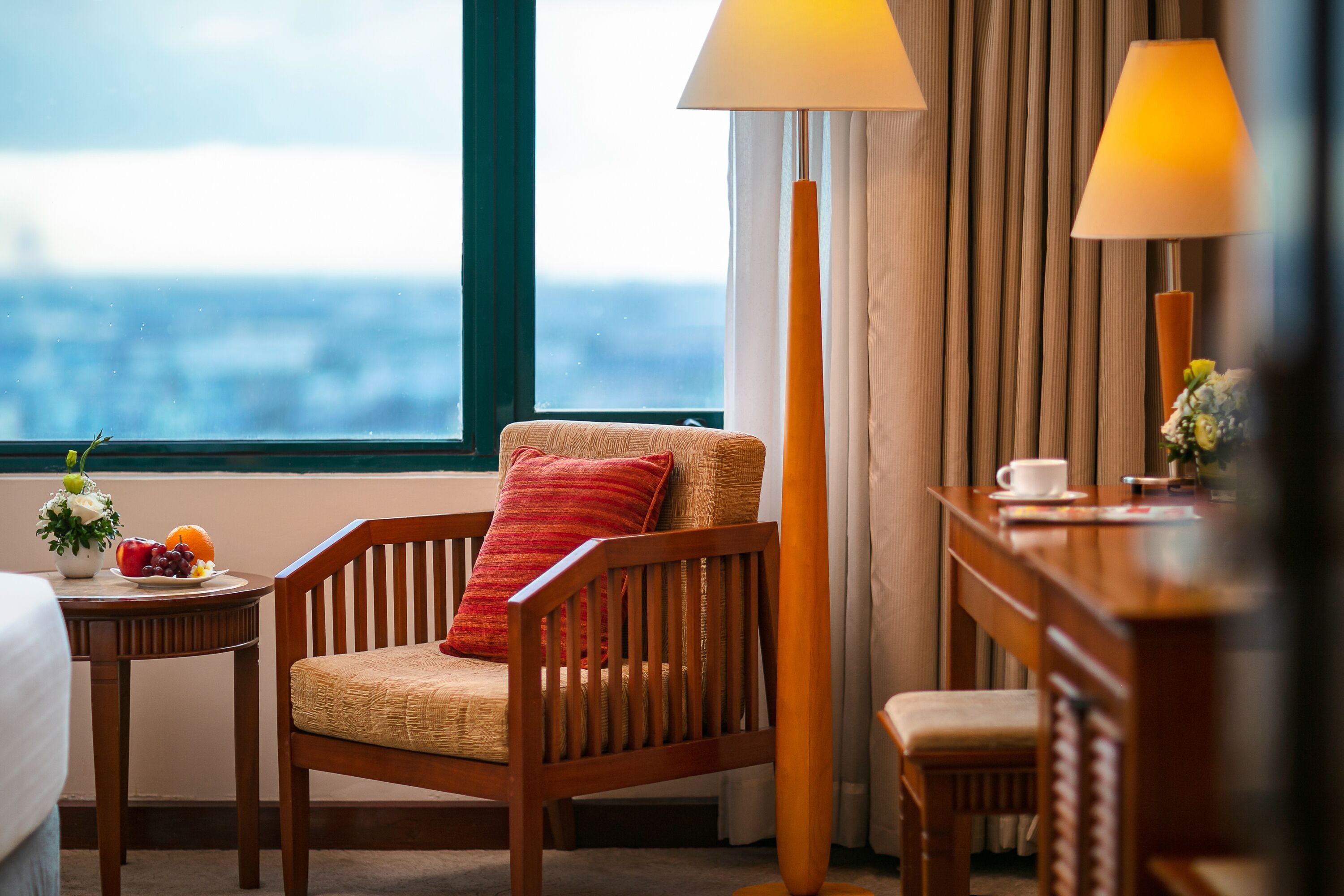 A hotel room with a wooden chair, a small table with fruit, a lamp, and a view of the city through a large window.