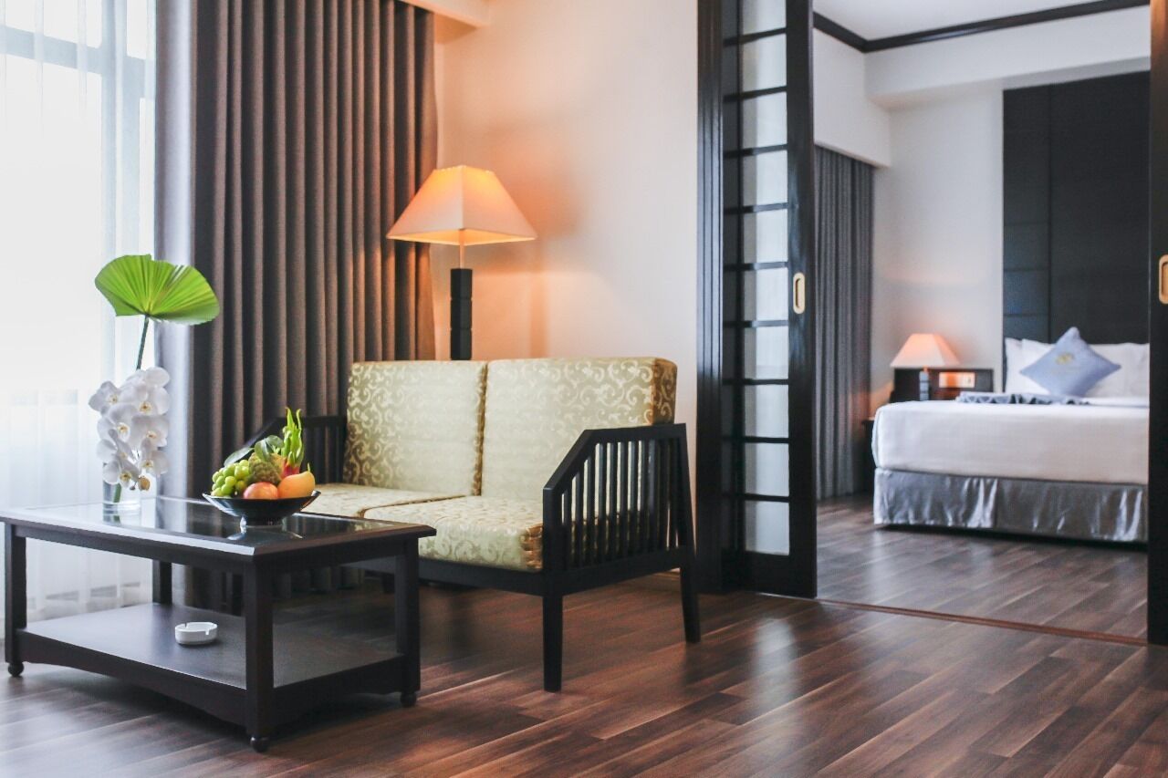 A hotel room with a sofa, a small table with a fruit bowl, a bed with pillows, and a nightstand with a lamp.