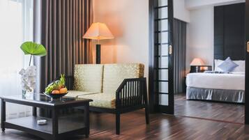 A hotel room with a sofa, a small table with a fruit bowl, a bed with pillows, and a nightstand with a lamp.