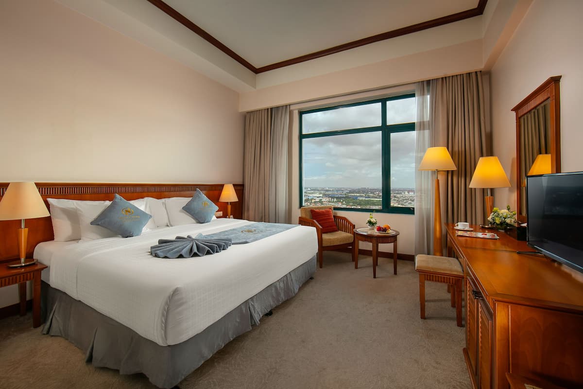 A hotel room with a large bed, a desk with a TV, a chair, and a view of the city through a large window.