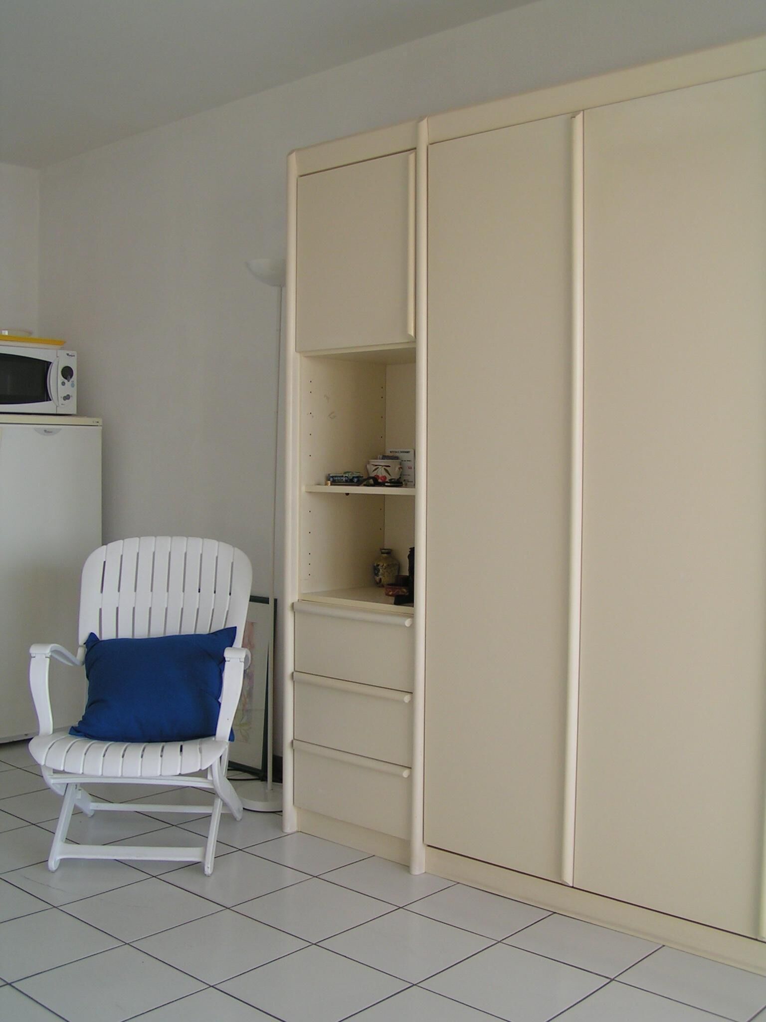 A white chair with a blue cushion, a white wardrobe with multiple shelves, and a microwave on top of a white cabinet.