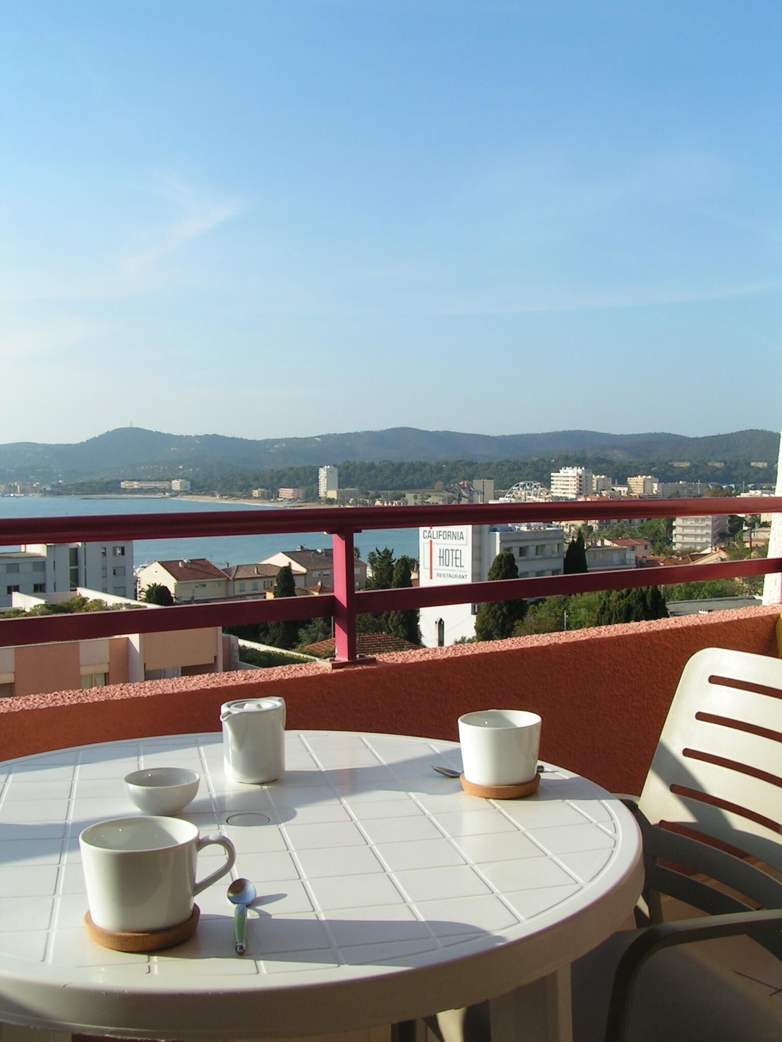 A round table with a white tablecloth and two white cups on coasters, a small decorative item, and a chair. In the background, there is a view of a cityscape with buildings and a hill.