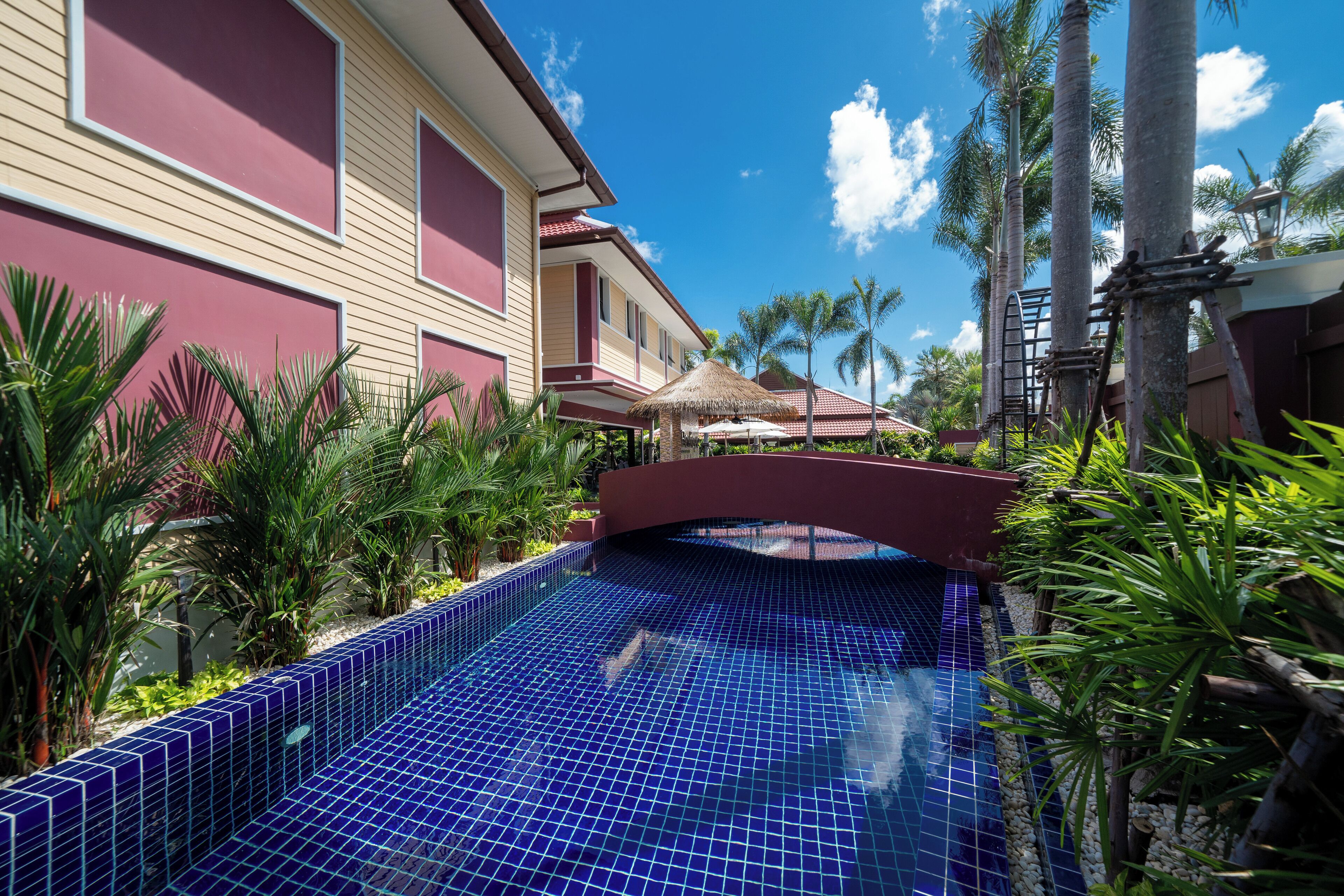 A swimming pool with blue tiles, surrounded by palm trees and a building with a red and beige facade.