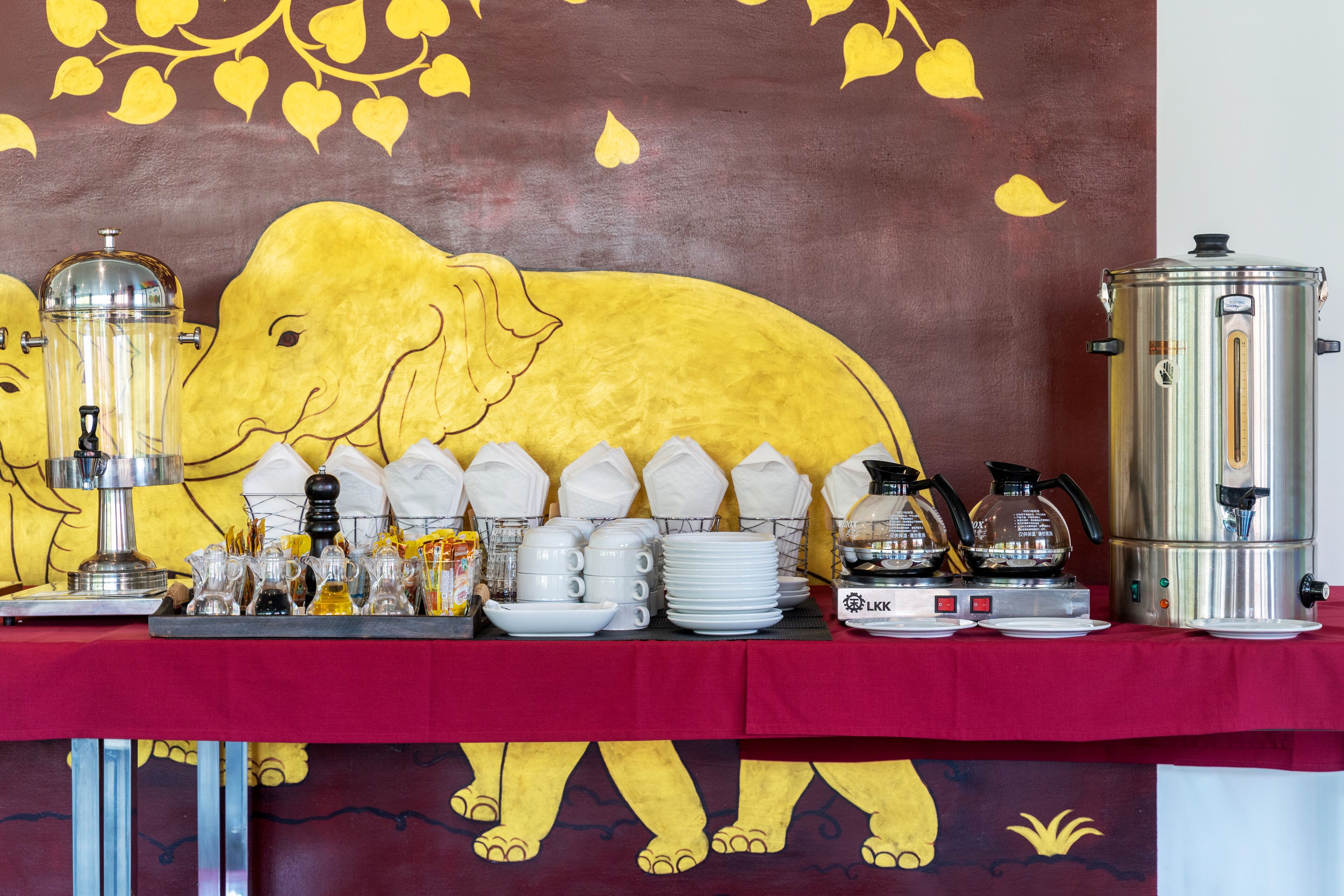 Una mesa de buffet con un gran mural de elefantes, una estación de café y diferentes platos y condimentos.