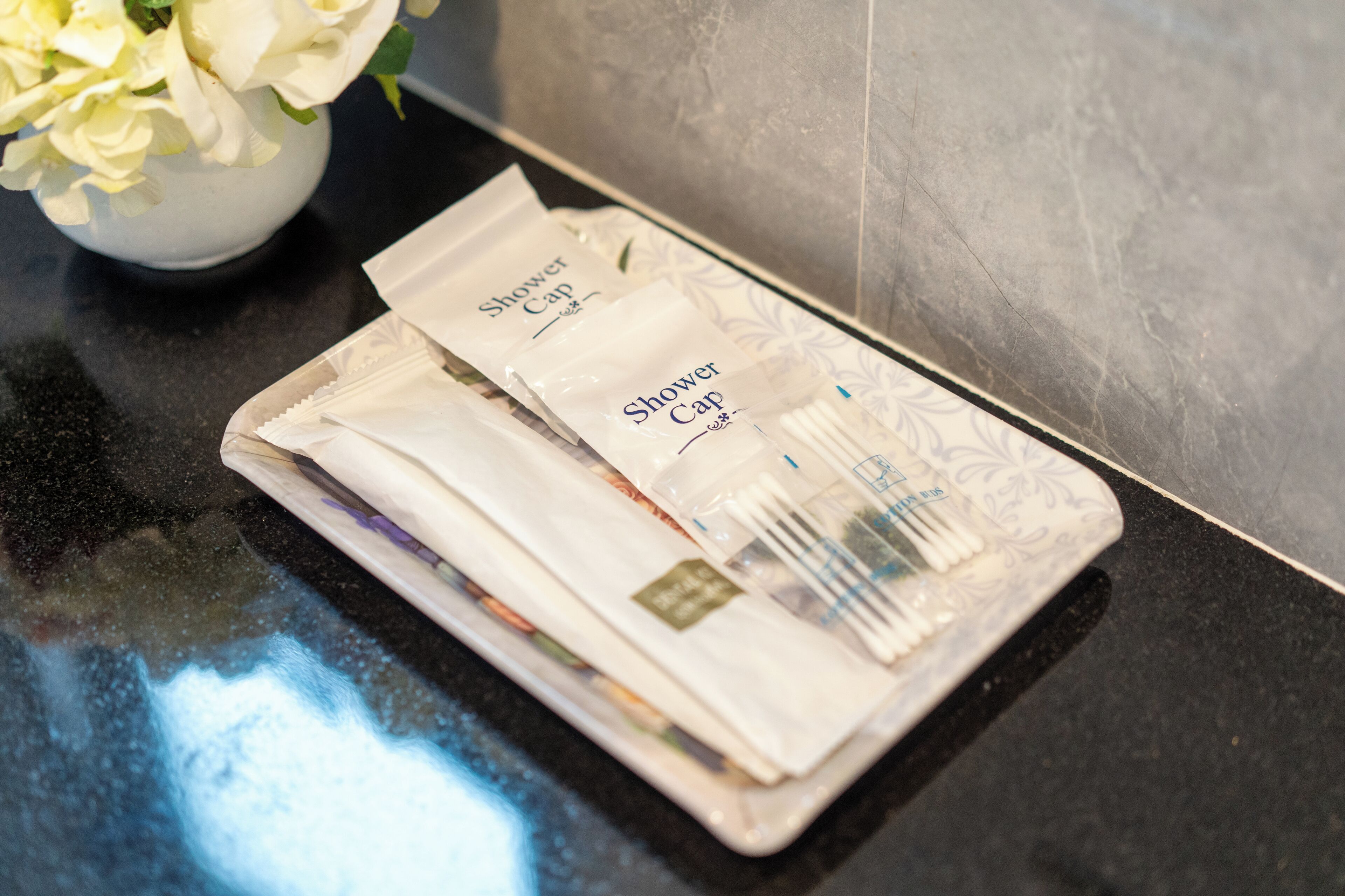 A bathroom counter with a white vase containing white flowers and several packages labeled "Shower Cap."