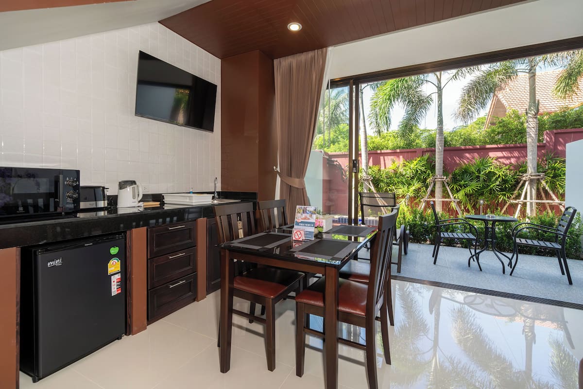 A compact kitchen with a refrigerator, microwave, and coffee maker, a dining table with chairs, and a view of a patio with plants and a red wall.