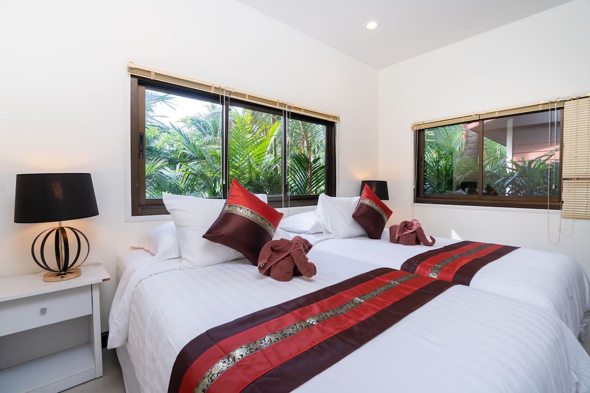 A hotel room with two beds, white bedding, red and brown pillows, a red and brown striped blanket, a bedside table with a lamp, and a window with a view of greenery.